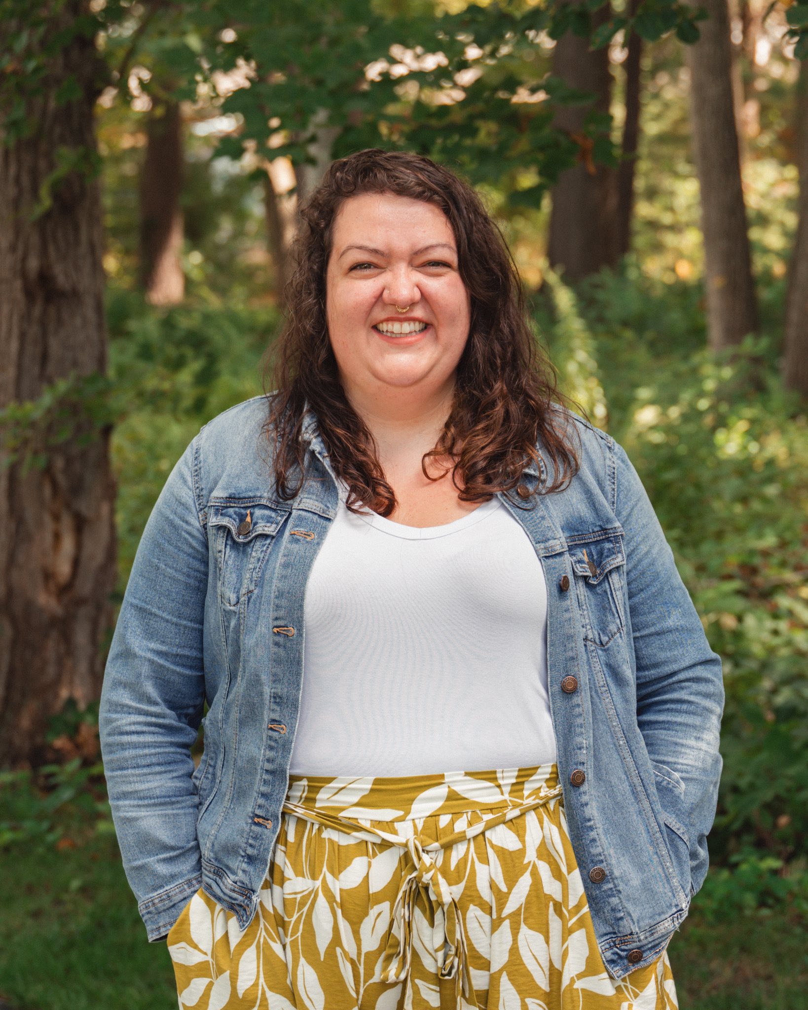 Shay is wearing a white shirt, green and white patterned pants tied in the front, and a blue denim jacket. She had brown curly hair and stands with her hands in her pockets outside in front of a wooded area.