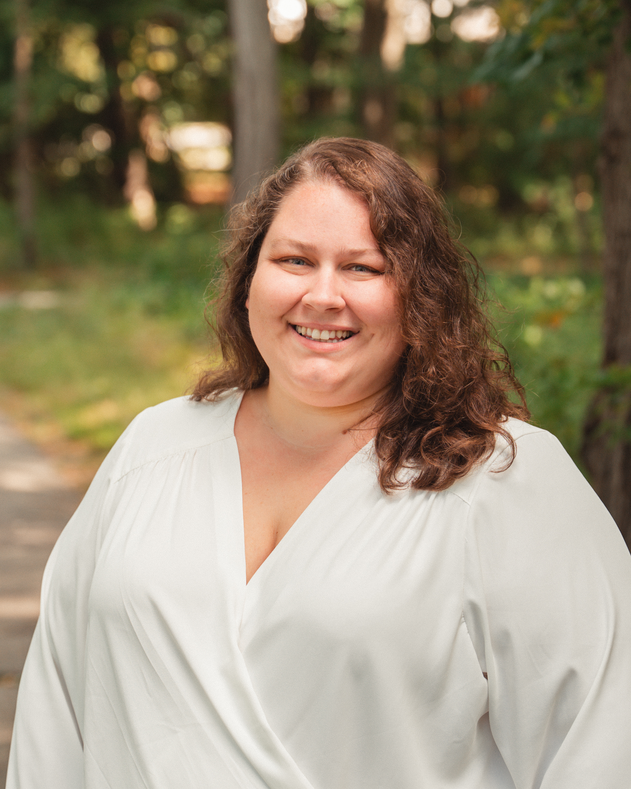 Cheryl Lawrence has wavy brown hair, wearing a white blouse smiling with a slight blurred outdoor background.