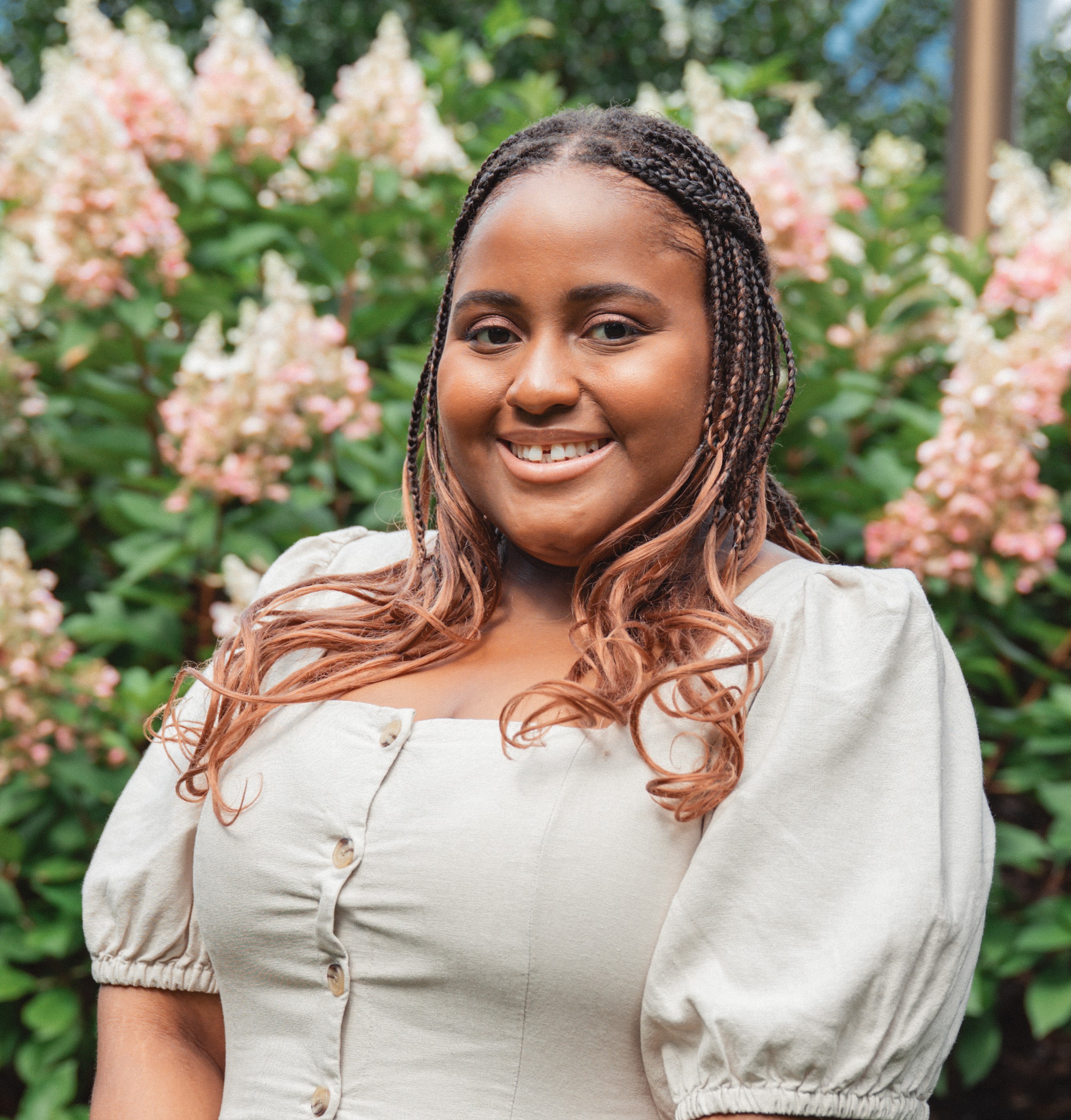 Tanisha has dark brown with light brown highlight braids, wearing beige dress and has a green bush with pink flowers background, smiling