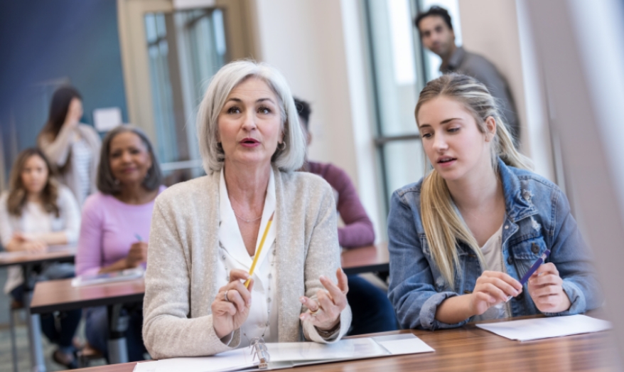 People sitting in a classroom setting, and the focus is on two people sitting a table with one person talking